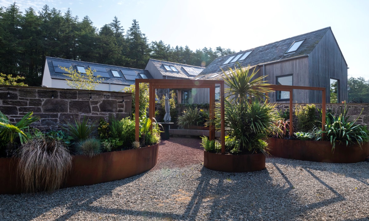 House made of collection of farm buildings with cladding and garden space with plants in foreground