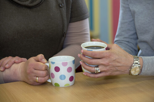 A close up photograph of two people at a table in conversation, cropped just below the neck to focus on their arms. Each person is holding a cup.