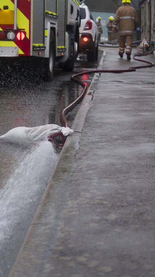 A photo of a wet street, with a fire engine in the distance. In the foreground is a hose which is ejecting water from a flooded property.