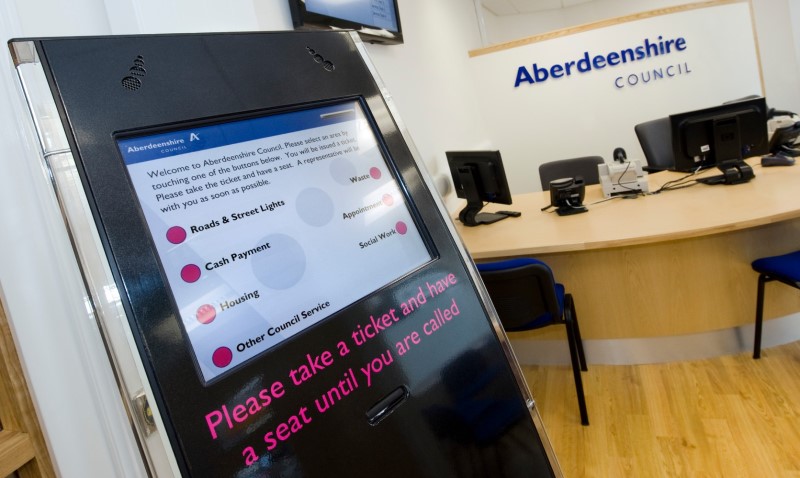 A digital computer screen at the Huntly service point with a reception table and chairs in the background below a sign which reads Aberdeenshire Council
