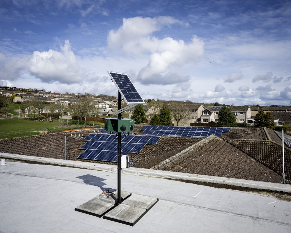 A solar-powered gull scarer on a metal stand on top of a school looking out over rooftops, houses and some trees