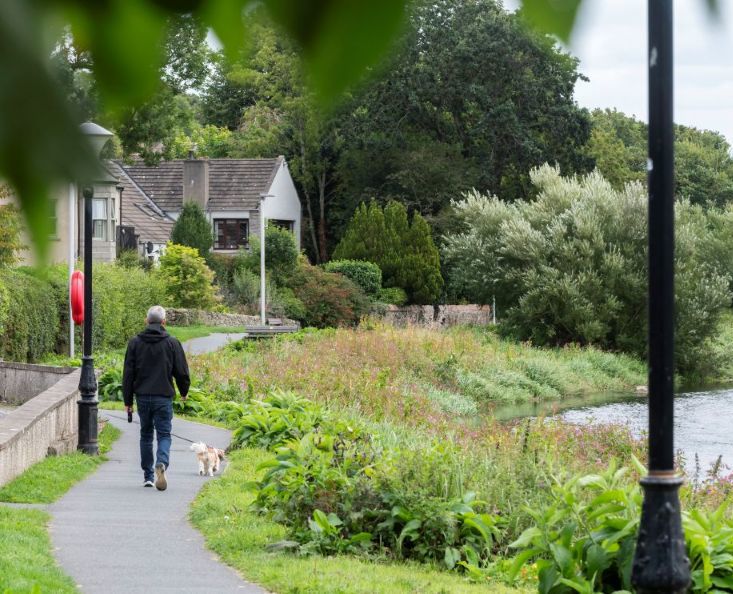 A man walking a dog along a path beside a river with houses and a wood in the background. 