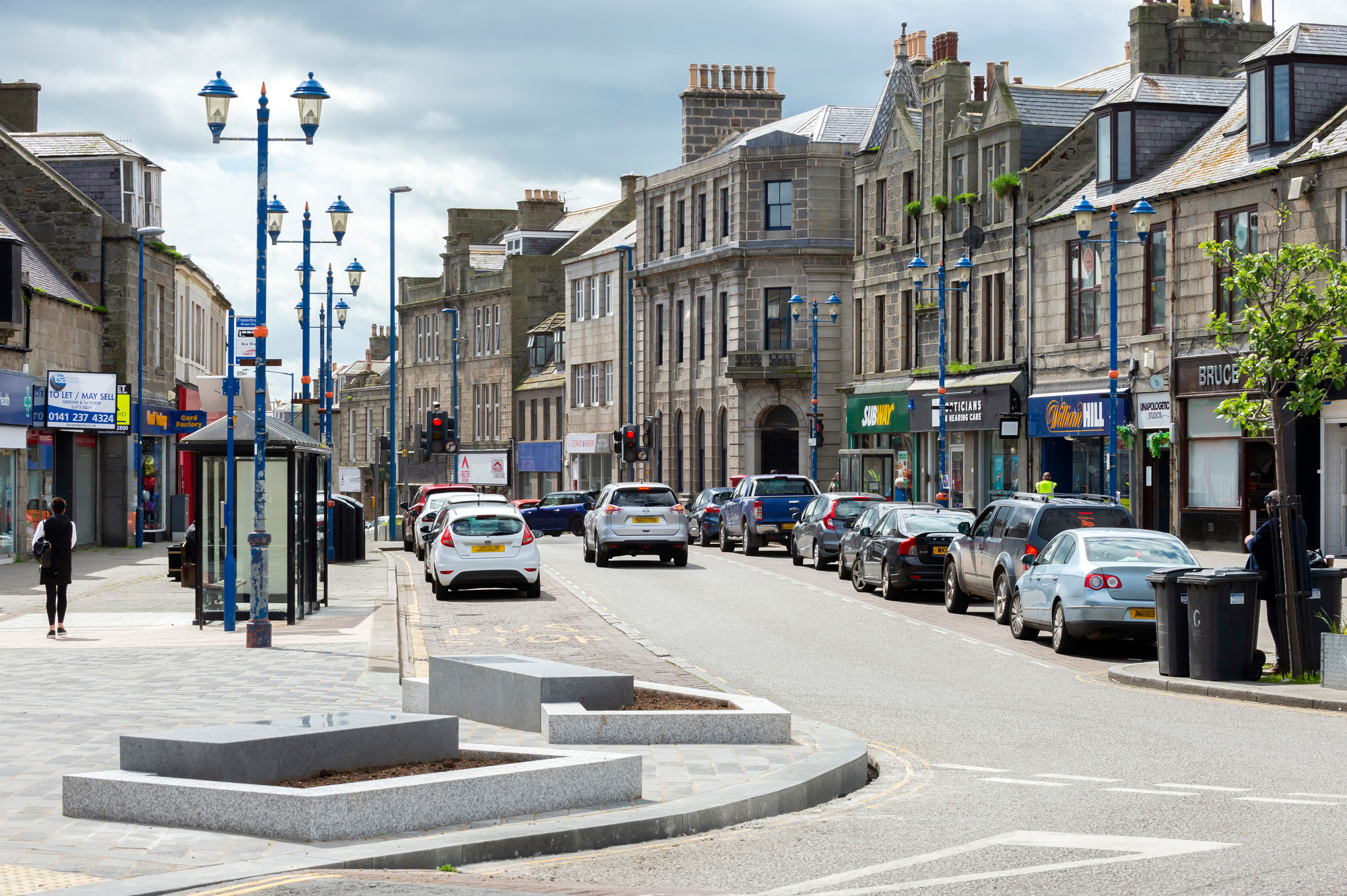 A photograph of Fraserburgh town centre featuring shops, cars, pedestrians, blue street lamps and some trees