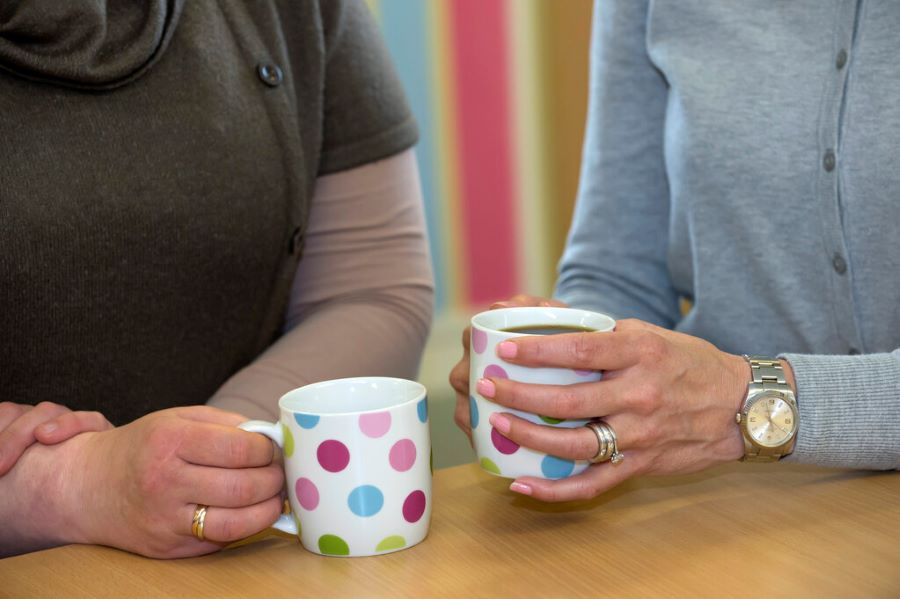 A close up of two people sat by a table, each holding a cup. The image shows their arms and torsos but not their faces. 